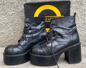 May include: A pair of black leather platform boots with laces and a chunky heel. The boots are sitting on a wooden surface.