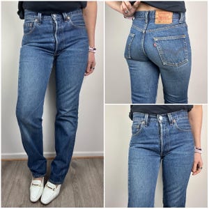 Op de afbeelding: Blauwe denim jeans met een klassiek vijf-zakken ontwerp. De jeans hebben een knoopsluiting en een rechte pijp. De achterkant heeft een rood Levi's-label en een klein zakje. De jeans worden gedragen met witte loafers.
