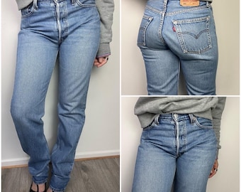 May include: Light blue denim jeans with a classic straight-leg fit. The jeans feature a button fly, five-pocket design, and a red Levi's tag on the back pocket. The jeans are worn with a gray sweatshirt and black shoes.