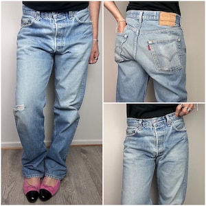 May include: Light blue denim jeans with a distressed knee. The jeans have a button fly and classic five-pocket design. A small red tag is visible on the back pocket. The jeans are worn with pink and black flats.
