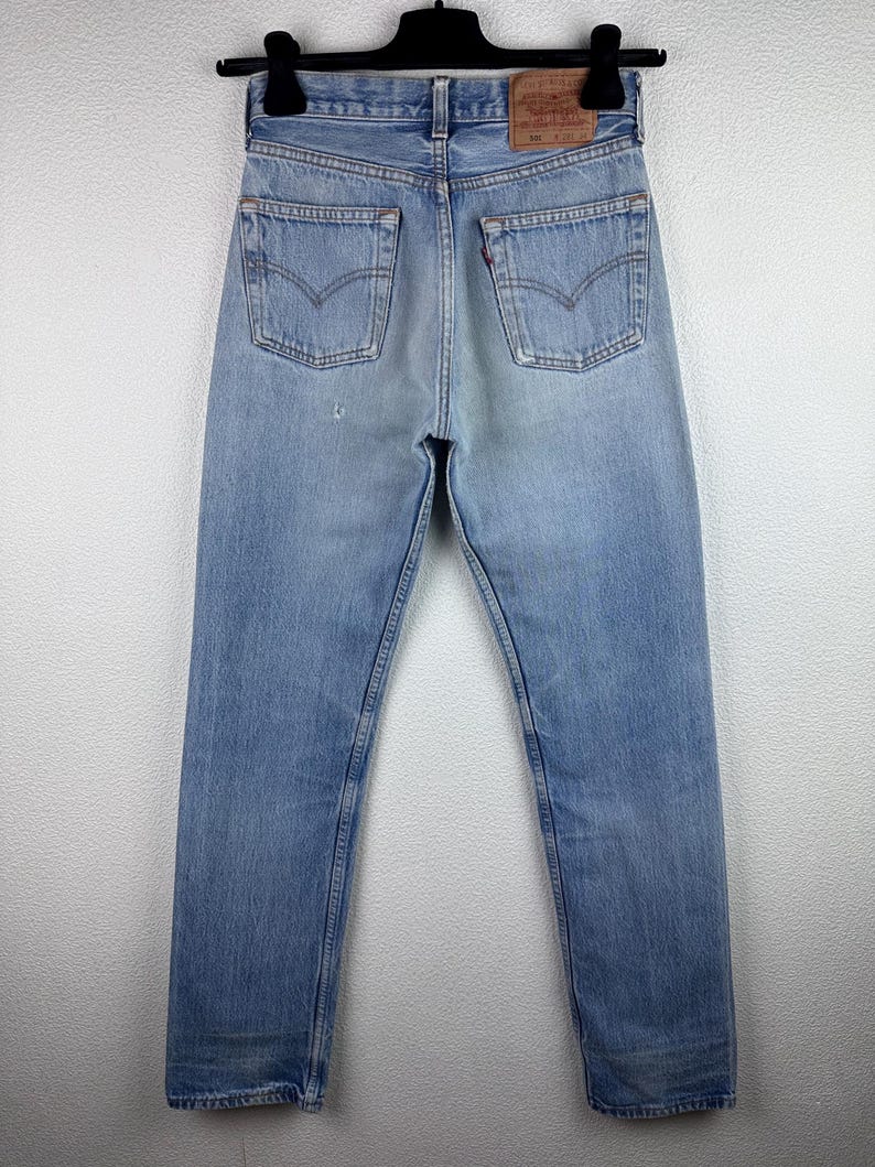 May include: Light blue denim jeans with classic five-pocket styling. The jeans feature a faded wash and a leather patch on the back. The jeans are hanging on a black hanger.