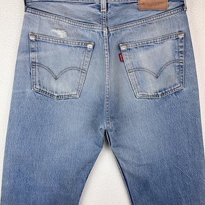 May include: Light blue denim jeans with a classic five-pocket design. The jeans feature a distressed look with faded areas and a small tear on the back pocket. A brown leather patch is visible at the waistband.