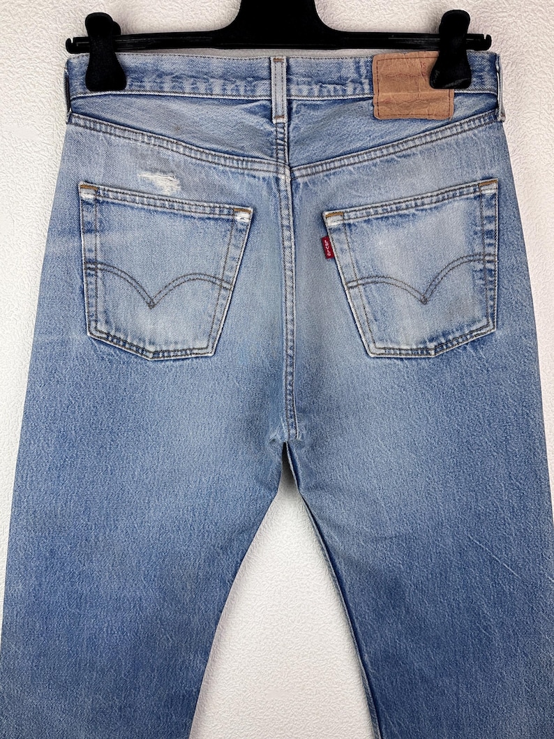 May include: Light blue denim jeans with a classic five-pocket design. The jeans feature a distressed look with faded areas and a small tear on the back pocket. A brown leather patch is visible at the waistband.