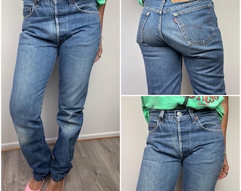 May include: Light blue denim jeans with a button fly and a classic five-pocket design. The jeans have a faded wash and a straight leg fit. The back features a leather patch and a red tab. The jeans are worn with a green t-shirt and pink flats.