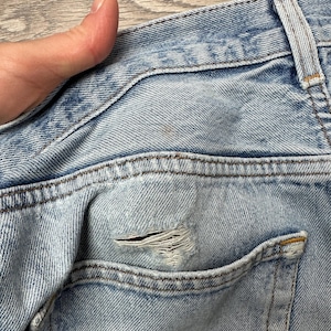 May include: Close-up of light blue denim jeans with a distressed pocket. The jeans show signs of wear and tear, with a visible hole in the pocket area. The stitching is visible, and the denim has a faded appearance.