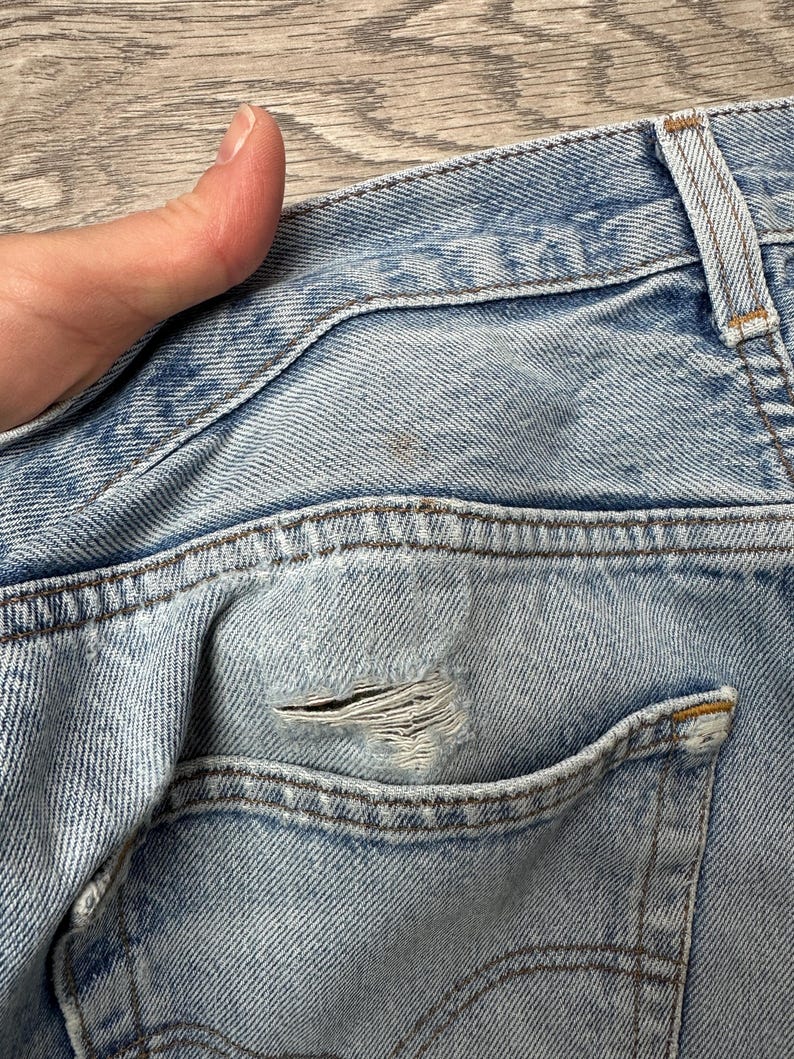 May include: Close-up of light blue denim jeans with a distressed pocket. The jeans show signs of wear and tear, with a visible hole in the pocket area. The stitching is visible, and the denim has a faded appearance.