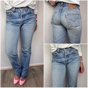 May include: Light blue denim jeans with a button fly and classic five-pocket design. The jeans have a faded wash and a straight leg fit. A small red tag is visible on the back pocket. The jeans are worn with pink flats.