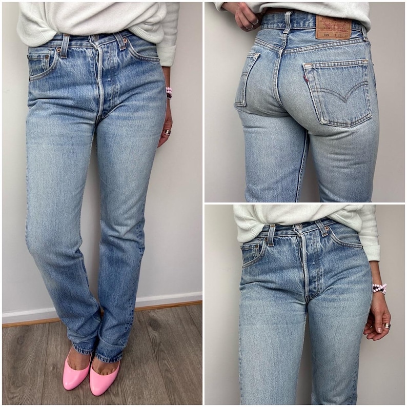 May include: Light blue denim jeans with a button fly and classic five-pocket design. The jeans have a faded wash and a straight leg fit. A small red tag is visible on the back pocket. The jeans are worn with pink flats.