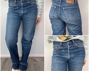 May include: Three-panel image of blue denim jeans. The jeans have a button fly, classic five-pocket design, and a faded wash. The back view shows a leather patch with red stitching. The jeans are worn with a gray sweatshirt.