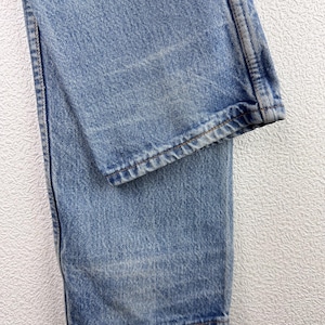 May include: Close-up of light blue denim jeans. The jeans show signs of wear and fading, with visible stitching along the seams and hem. The fabric has a textured appearance, and the overall look is casual and classic.