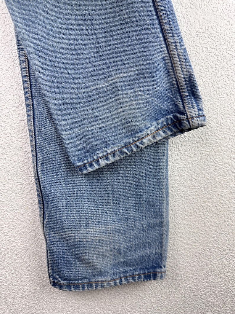 May include: Close-up of light blue denim jeans. The jeans show signs of wear and fading, with visible stitching along the seams and hem. The fabric has a textured appearance, and the overall look is casual and classic.