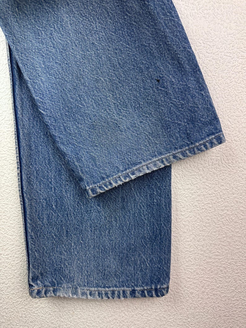 May include: Close-up of light blue denim jeans. The jeans are folded, showing the texture of the fabric and the hem of the leg. The denim has a classic, worn look.