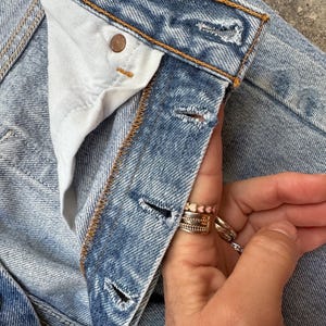 May include: Close-up of light blue denim jeans. The jeans have a button closure and a button on the waistband. The stitching is a contrasting orange color. The jeans are worn and have a faded appearance. The fly is unbuttoned.