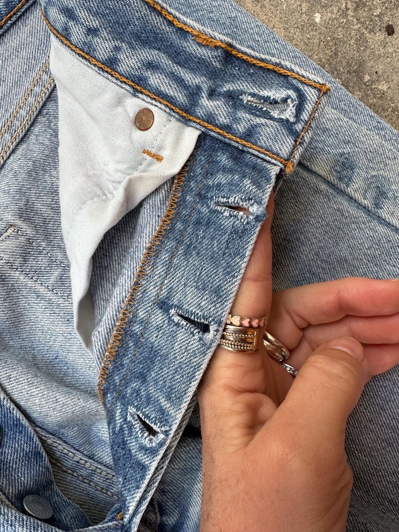 May include: Close-up of light blue denim jeans. The jeans have a button closure and a button on the waistband. The stitching is a contrasting orange color. The jeans are worn and have a faded appearance. The fly is unbuttoned.