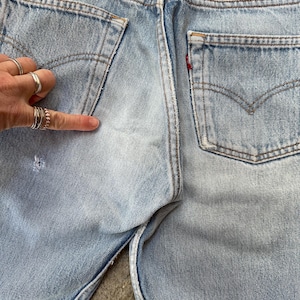 May include: Light blue denim jeans with classic five-pocket design. Features include a faded wash, distressed details, and a red tab on the back pocket. The jeans have a button fly and a leather patch.