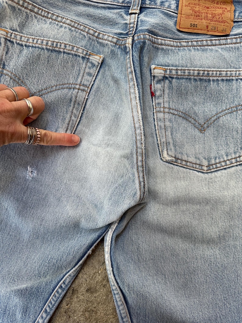 May include: Light blue denim jeans with classic five-pocket design. Features include a faded wash, distressed details, and a red tab on the back pocket. The jeans have a button fly and a leather patch.