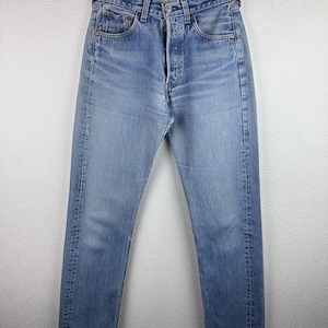 May include: Light blue denim jeans with a button fly and classic five-pocket design. The jeans are hung on a black hanger against a white wall. The denim has a faded appearance.