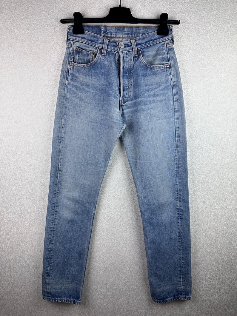 May include: Light blue denim jeans with a button fly and classic five-pocket design. The jeans are hung on a black hanger against a white wall. The denim has a faded appearance.
