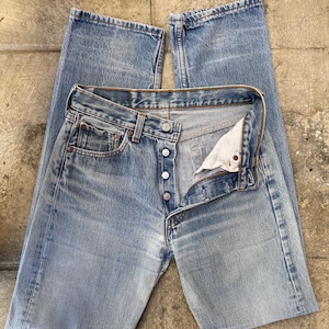 May include: Light blue denim jeans with a button fly, open to reveal the interior. The jeans have a classic five-pocket design and a faded wash. The jeans are laid flat on a textured surface.
