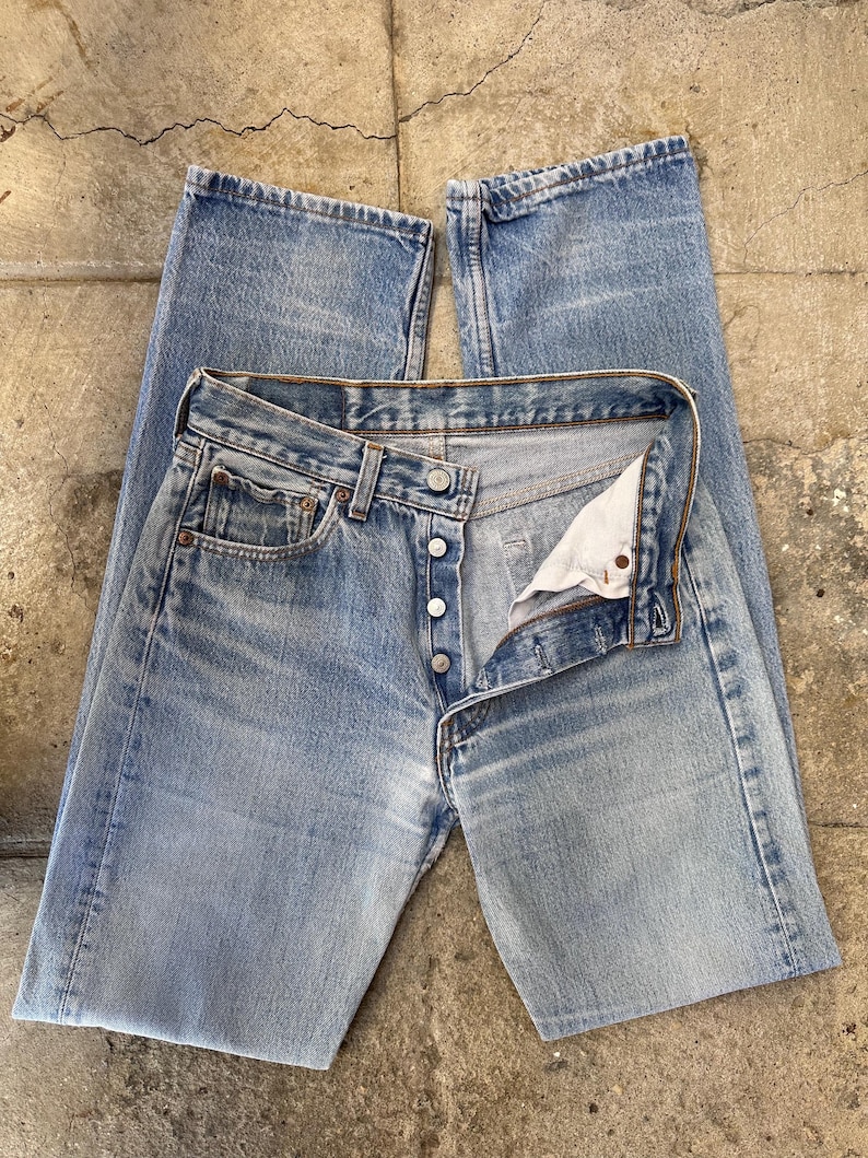 May include: Light blue denim jeans with a button fly, open to reveal the interior. The jeans have a classic five-pocket design and a faded wash. The jeans are laid flat on a textured surface.