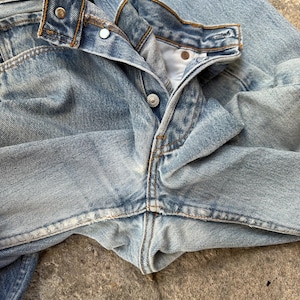 May include: Close-up of light blue denim jeans. The jeans are unbuttoned, revealing the button and buttonhole. The metal rivets and stitching are visible. The denim has a faded, worn appearance.