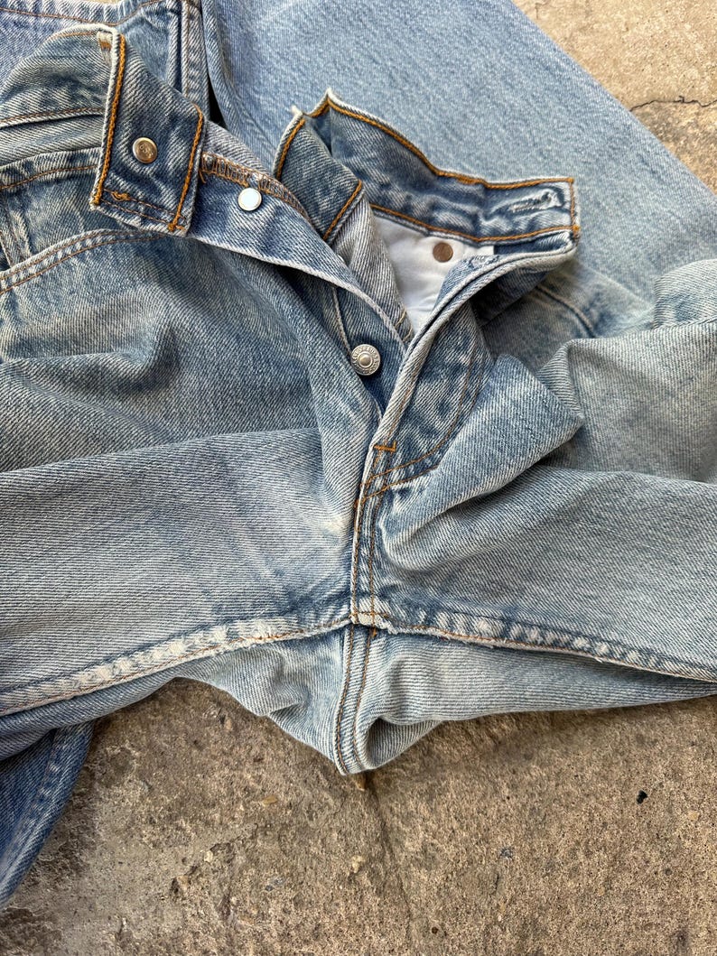 May include: Close-up of light blue denim jeans. The jeans are unbuttoned, revealing the button and buttonhole. The metal rivets and stitching are visible. The denim has a faded, worn appearance.