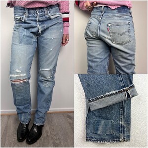 Op de afbeelding: Lichtblauwe denim jeans met distressed details, waaronder gescheurde knieën en gerepareerde plekken. De jeans heeft een knoopsluiting en een klassiek vijf-zakken ontwerp. De manchetten zijn opgerold.