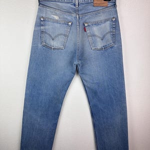 May include: Light blue denim jeans with a classic five-pocket design, visible back pockets, and a leather patch. The jeans are hanging on a black hanger against a white background. The jeans have a distressed look.