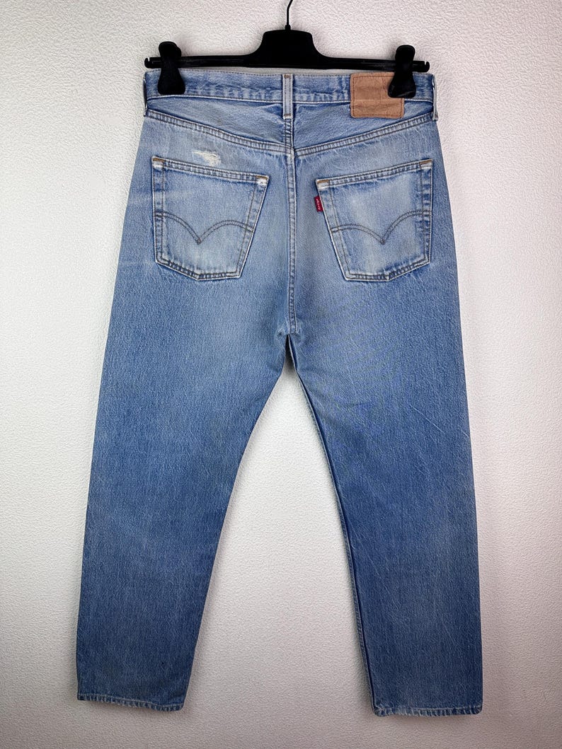 May include: Light blue denim jeans with a classic five-pocket design, visible back pockets, and a leather patch. The jeans are hanging on a black hanger against a white background. The jeans have a distressed look.