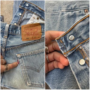 May include: Close-up of light blue denim jeans. The jeans feature a classic five-pocket design, a red tab on the back pocket, and a brown leather Levi Strauss & Co. label. The jeans have metal button closures and copper stitching.