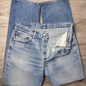 May include: Light blue denim jeans with a button fly, classic five-pocket design, and a faded wash. The jeans are laid flat, showcasing the details of the fabric and construction. The denim has a worn appearance.