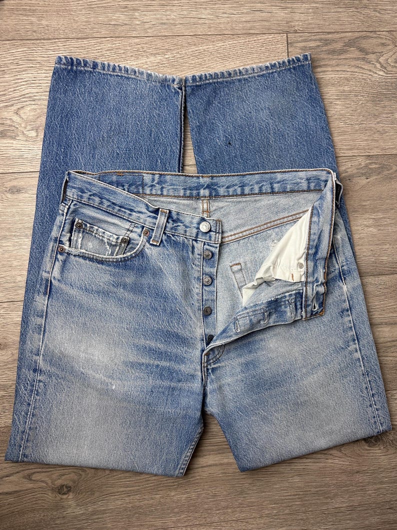 May include: Light blue denim jeans with a button fly, classic five-pocket design, and a faded wash. The jeans are laid flat, showcasing the details of the fabric and construction. The denim has a worn appearance.
