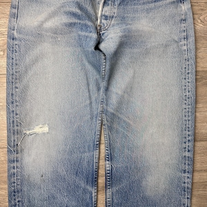 May include: Faded blue denim jeans with a button fly and distressed details. The jeans have a worn appearance with a tear on the left leg. The denim is a light wash with visible fading and wear.