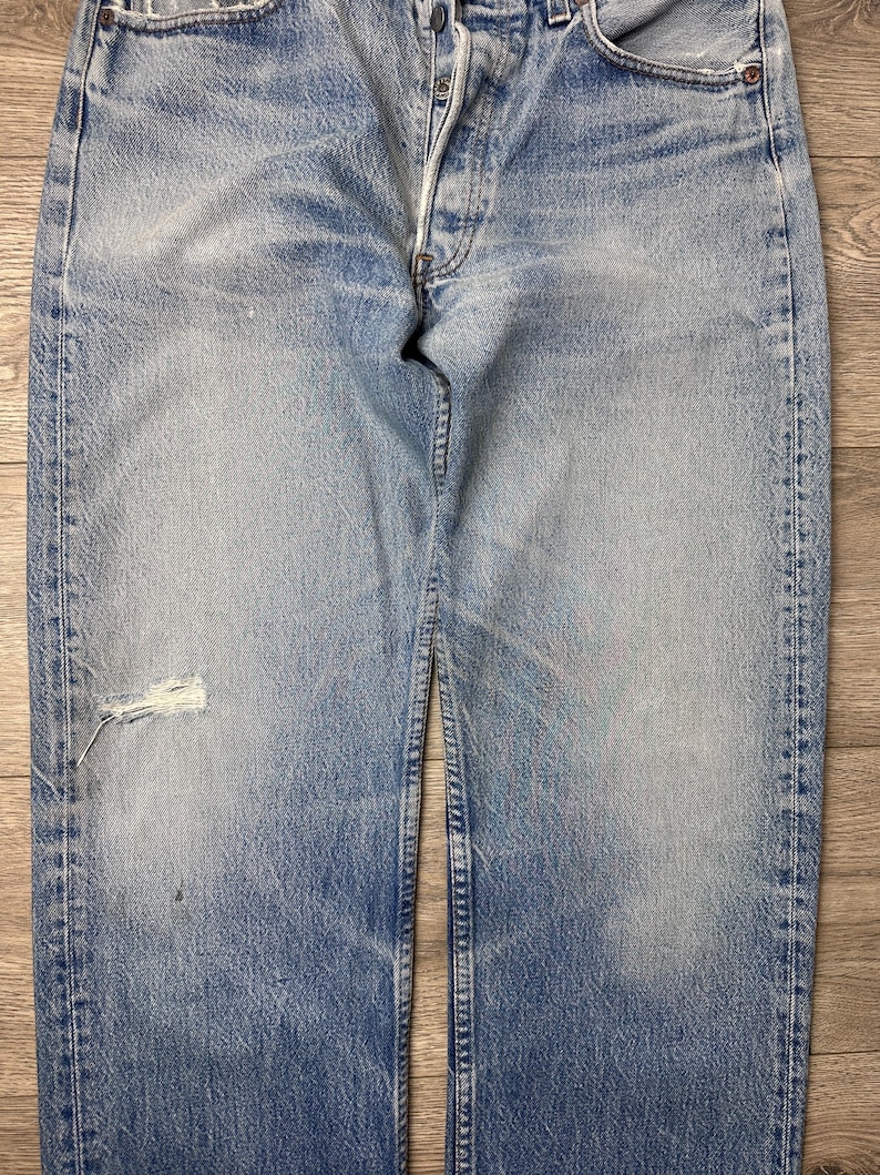 May include: Faded blue denim jeans with a button fly and distressed details. The jeans have a worn appearance with a tear on the left leg. The denim is a light wash with visible fading and wear.