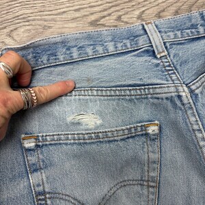 May include: Close-up of light blue denim jeans, showing the back pocket and waistband. The jeans have a distressed look with a small tear near the pocket. The pocket has a classic curved design with a brown stitch. The jeans are faded and worn.