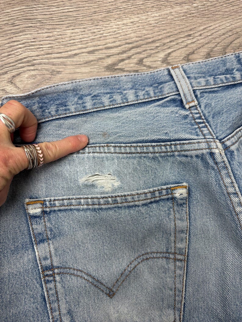 May include: Close-up of light blue denim jeans, showing the back pocket and waistband. The jeans have a distressed look with a small tear near the pocket. The pocket has a classic curved design with a brown stitch. The jeans are faded and worn.