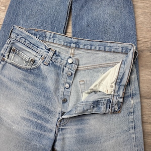 May include: Light blue denim jeans with a button fly and classic five-pocket design. The jeans show signs of wear and fading, with a slightly distressed look. The jeans are laid flat, showcasing the front and partially open fly.