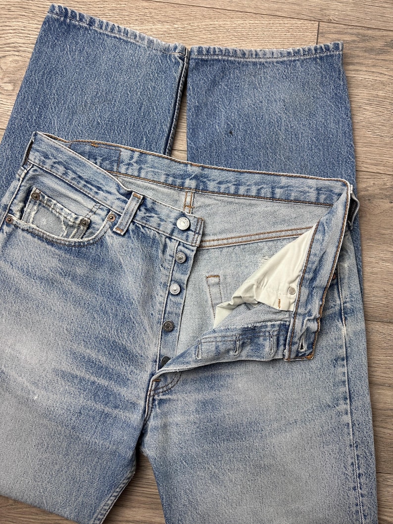 May include: Light blue denim jeans with a button fly and classic five-pocket design. The jeans show signs of wear and fading, with a slightly distressed look. The jeans are laid flat, showcasing the front and partially open fly.