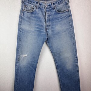 May include: Light blue denim jeans with a button fly and a distressed knee. The jeans are faded and worn, with visible wear marks. The jeans are hanging on a black hanger against a white background.