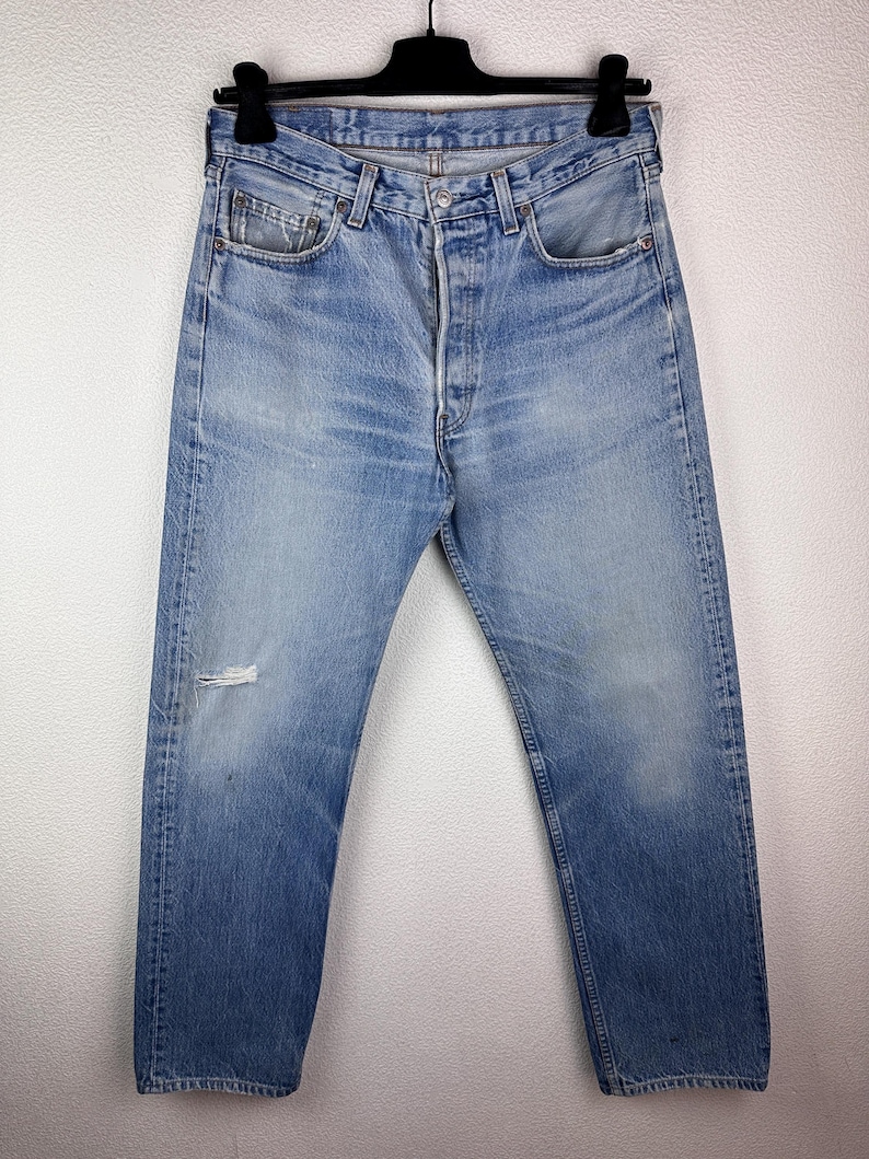 May include: Light blue denim jeans with a button fly and a distressed knee. The jeans are faded and worn, with visible wear marks. The jeans are hanging on a black hanger against a white background.