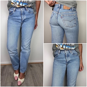 May include: Light blue high-waisted denim jeans featuring a button fly closure, five-pocket styling, and a tan leather patch on the back waistband. The jeans have a straight-leg fit with a classic medium wash finish and a red tab on the back pocket.