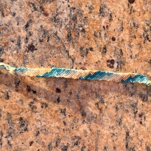 May include: A blue, yellow, and white friendship bracelet made with string. The bracelet is lying on a brown, speckled surface.