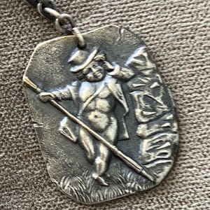 May include: A rectangular silver-toned metal pendant featuring a raised relief of a figure wearing a top hat and holding a long staff. The figure stands in a grassy area, and the metal has an oxidized finish with dark recessed details.