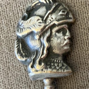 May include: A silver-toned metal spoon handle finial featuring a detailed relief sculpture of a profile view of a person wearing a plumed helmet and armor. The design includes textured hair, a visor, and a scale-patterned chest piece on the figure.