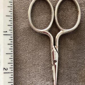 May include: Small silver-colored metal scissors with oval finger loops and a pointed tip. The blades are marked with the word GERMANY. A white ruler is positioned to the left, showing the scissors measure approximately 2.5 inches in length.