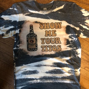 以下が含まれることがあります： グレーのタイダイTシャツに、漂白されたデザインが施されています。デザインには、ティトスの手作りウォッカのボトルと「Show Me Your Titos」というテキストが描かれています。