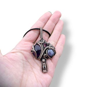 May include: A copper wire wrapped pendant featuring three purple and grey gemstones. The pendant is attached to a black cord.