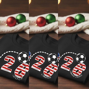 May include: Black t-shirts with a soccer ball design and the number "26" in red and white stripes, with the text "U.S.A.", "America", and "United States" below. The shirts are displayed with red and green ornaments.
