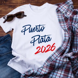 以下が含まれることがあります： 青と赤の筆記体で「Puerto Plata 2026」と書かれた白いTシャツ。シャツは、ブルージーンズ、チェック柄のシャツ、サングラスと組み合わせています。アイテムは木製の表面に配置されています。