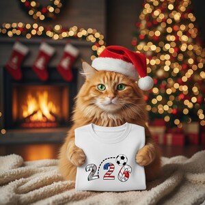 Puede incluir: Un gato jengibre con un gorro de Papá Noel sostiene una camiseta blanca con los números "226" y un gráfico de balón de fútbol. El "2" es plateado, el segundo "2" tiene rayas rojas y azules, y el "6" es rojo y blanco. Un árbol de Navidad y una chimenea están en el fondo.